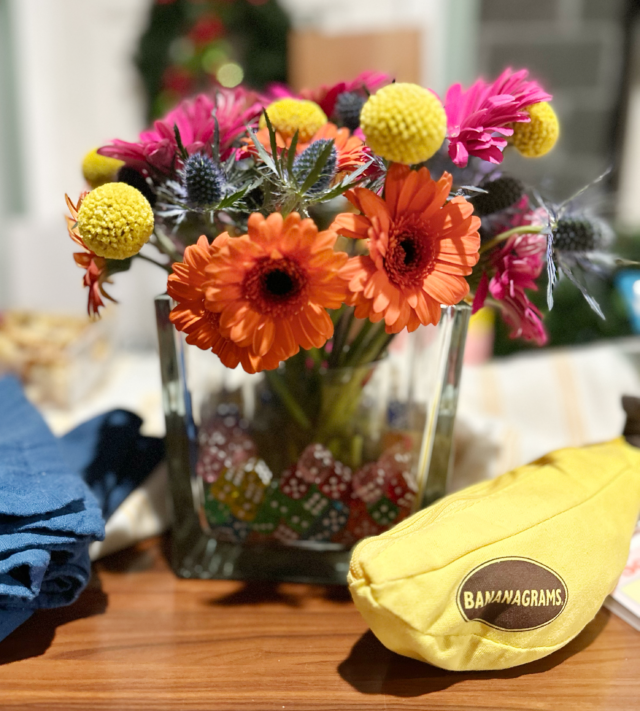 bightly colored flowers in a clear vase that is filled with colorful dice. Bananagrams game is on the table