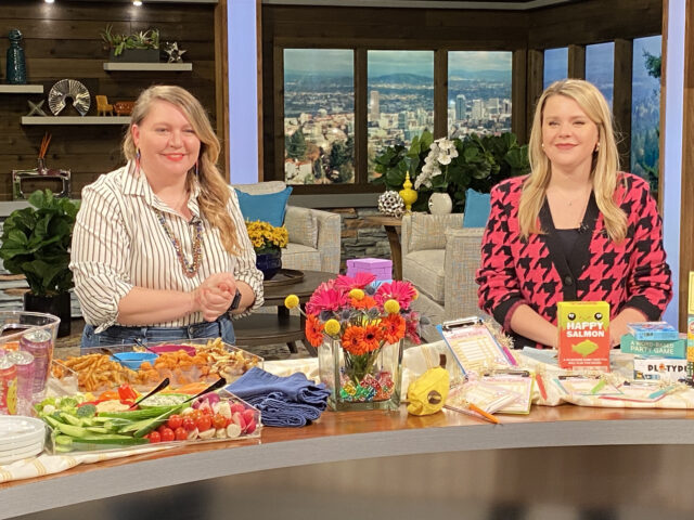 Two women standing behind a counter with board games, a game related centerpiece, and food boards. 