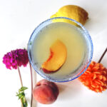 Cocktail overhead shot with peach slice in it. Peach and lemon on table with brightly colored mum flowers.