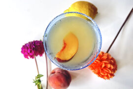 Cocktail overhead shot with peach slice in it. Peach and lemon on table with brightly colored mum flowers.