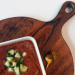 bowl of chilled vegetable soup with cucumbers on brown cutting board with gold spoon