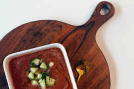 bowl of chilled vegetable soup with cucumbers on brown cutting board with gold spoon