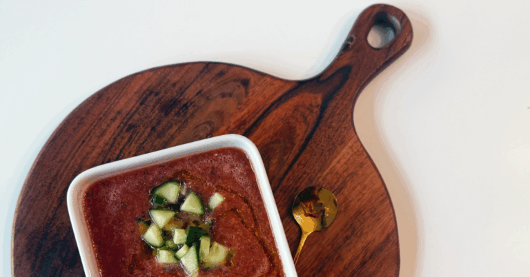 bowl of chilled vegetable soup with cucumbers on brown cutting board with gold spoon